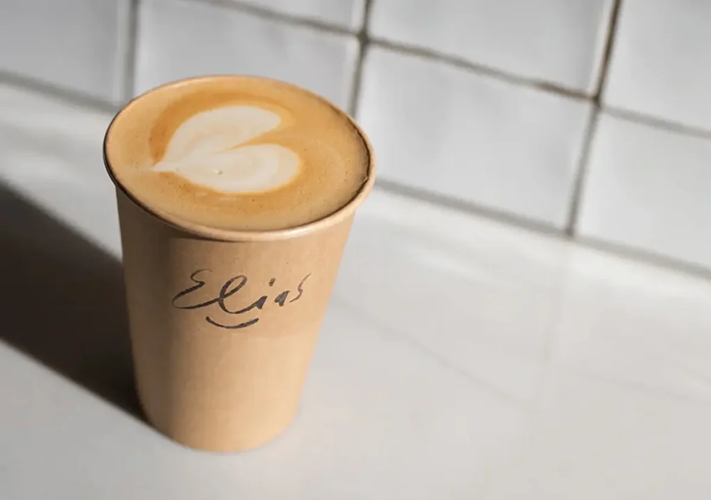 A paper cup of coffee with heart-shaped latte art sits on a counter; the name "Elias" is written on the cup in black marker, making this food and drinks moment extra personal.